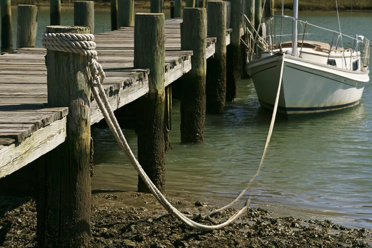 Low tide at a fixed dock: a dock line wrapped around the top of a piling droops across the exposed mud to a moored sailboat